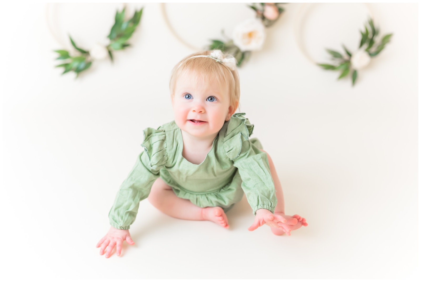 1 year old in green romper smiling at camera, studio session, organic floral hoops in background